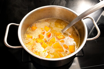 Cooking a vegetarian pumpkin soup from Hokkaido or red kuri squash in a steel pot on a black stove,...