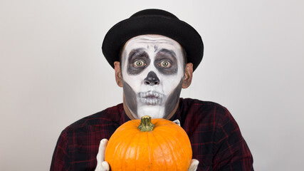 A scary clown looks at the camera, holds a pumpkin in his hands and threatens her with a knife. Halloween costume.