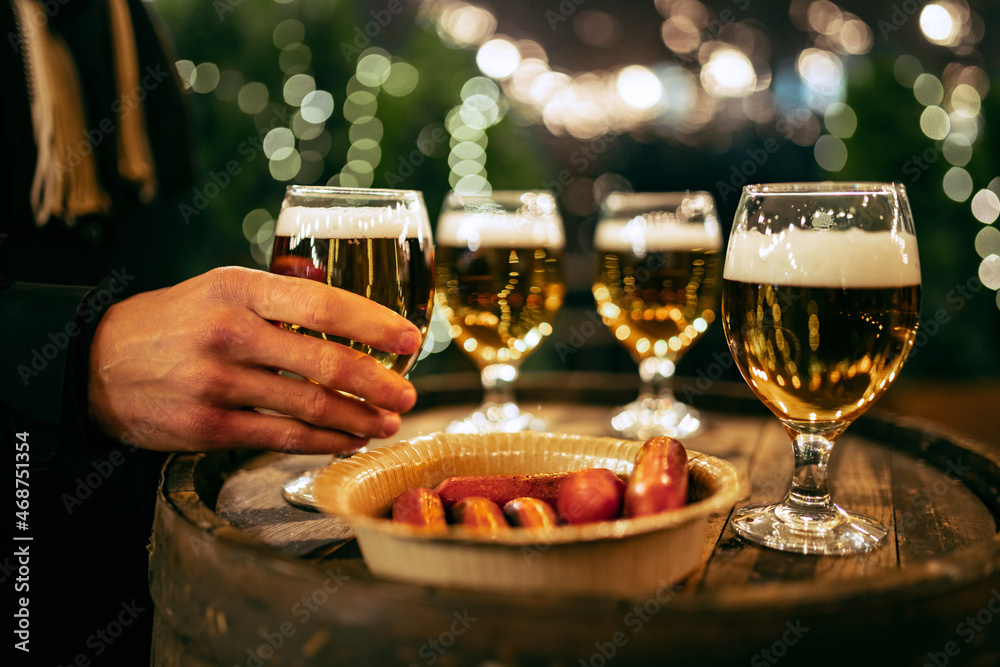 Wall mural Male hand holding glass of light frothy beer and sausages on wooden old barrel roll. Concept of drinks, fun, meeting, oktoberfest, winter fair