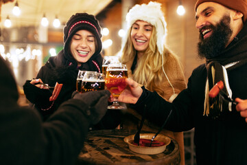 Close-up happy smiling friends with cups of mulled wine spending time together at winter fair at...