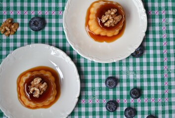 Flans with a walnut on top on a green and white checkered tablecloth surrounded by blueberries