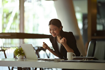 Fototapeta premium Photo of a businesswoman making a winner gesture while standing at the smart working desk over the comfortable office as a background.