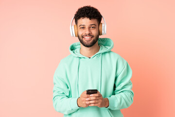 Young Moroccan man isolated on pink background listening music with a mobile and looking front