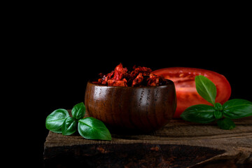 Diced dried tomato in the wooden bowl, basil leaves and plum tomato half on the wooden board against the black background. Natural ingredient