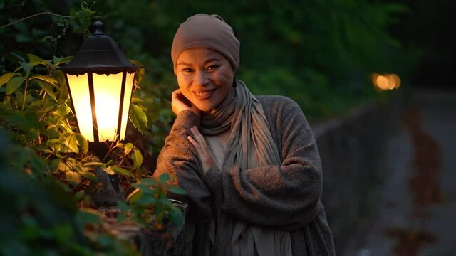 Asian woman stands smiling in the autumn evening in park. In the light of the soffit of the lantern and Fascinatingly looks into camera. Slow motion