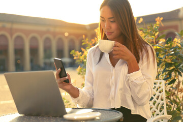 Self employed young woman holding a cup of coffee and smart phone working with her laptop in an outdoor cafe. - 468756719