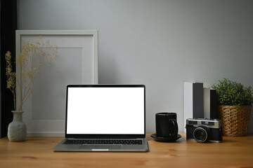 Photo of a white blank screen computer laptop on the wooden table surrounded by a coffee cup, vintage camera, empty picture frame potted plant and books.