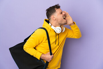 Young sport caucasian man with bag isolated purple background shouting with mouth wide open to the side