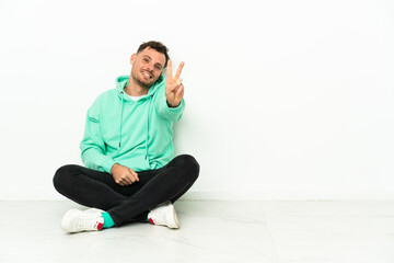 Young handsome caucasian man sitting on the floor smiling and showing victory sign