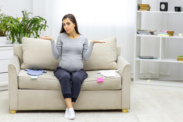 Fototapeta premium Photo of pregnant woman sitting on the sofa with child clothes.