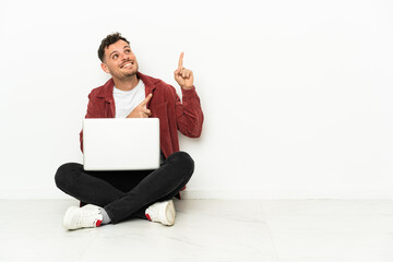 Young handsome caucasian man sit-in on the floor with laptop pointing with the index finger a great idea