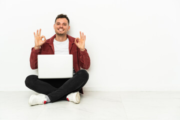 Young handsome caucasian man sit-in on the floor with laptop showing an ok sign with fingers