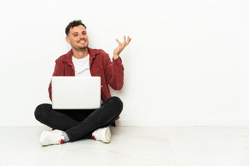 Young handsome caucasian man sit-in on the floor with laptop extending hands to the side for inviting to come