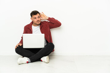 Young handsome caucasian man sit-in on the floor with laptop listening to something by putting hand on the ear