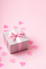 Silver glitter gift box with pink ribbon bow on pink background with confetti.