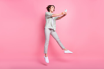 Fototapeta na wymiar Photo of sweet impressed young woman dressed beige clothes jumping high holding modern device isolated pink color background