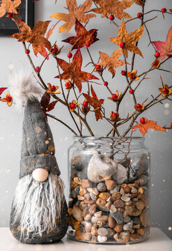 Christmas dwarf sitting with autumn branches in glass jar with pebbles grey minimalistic background holiday winter spirit