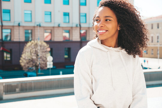 Beautiful black woman with afro curls hairstyle.Smiling hipster model in white hoodie. Sexy carefree female posing on the street background. Cheerful and happy outdoors