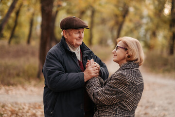 Elderly man looking at his elegant retired woman with love, warming her hands and enjoying her beauty, senior married couple dancing in autumn park, celebrating anniversary, affection