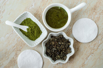 Three small white bowls with dry green tea leaves, green tea face or hair mask and matcha powder and cotton pads. Natural beauty treatment, skin care concept. Top view, copy space.