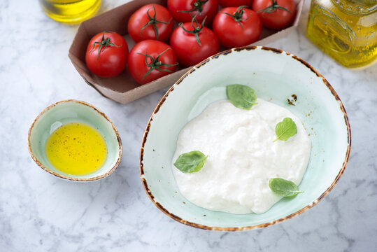 Green bowl with stracciatella di bufala, olive oil and red tomatoes over light-grey marble background, high angle view, horizontal shot