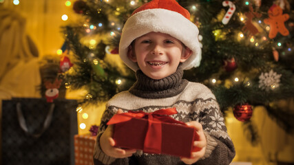 Fototapeta na wymiar Portrait of smiling boy in Santa hat holding Christmas present box and smiling in camera. Families and children celebrating winter holidays.