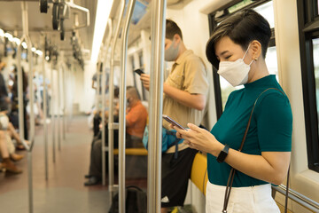 A beautiful smart look asian woman with disposable face mask on a city public train using her smartphone while travel to office. Urban, New normal, Lifestyle, Technology, Covid 19, Pandemic, 5G, FOMO.