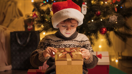 Fototapeta na wymiar Portrait of amazed and excited boy in Santa hat looking inside Christmas present box. Families and children celebrating winter holidays.