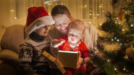 Fototapeta na wymiar Happy family with children sitting in armchair at Christmas tree and looking inside glowing Christmas present box. Families and children celebrating winter holidays.