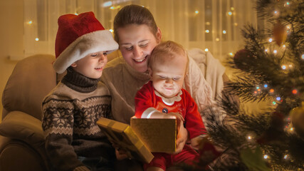 Fototapeta na wymiar Cute baby boy sitting on mothers lap in armchair and looking inside glowing Christmas present box. Families and children celebrating winter holidays.