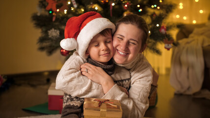 Fototapeta na wymiar Portrait of happy smiling mom and son in Santa hat hugging next to Christmas tree at house