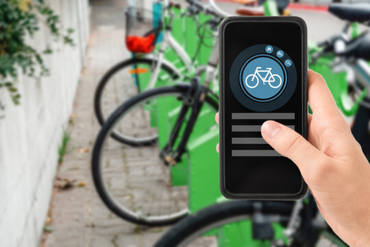 technology, transport and sustainability concept - close up of hand holding smartphone with mobile app and bicycle icon on screen over electric bike parking and charging station in city on background