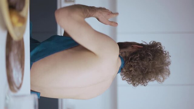 Young sexy guy is spinning on chair. And sends kisses and shows heart. Curly young man in denim blue apron having breakfast in morning light