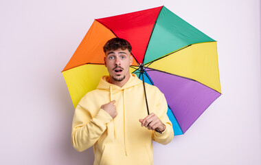 young handsome man looking shocked and surprised with mouth wide open, pointing to self. umbrella...