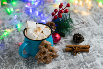 Obraz na płótnie Canvas Delicious cocoa with cinnamon and marshmallows in a blue cup, star anise, cinnamon sticks, Christmas decor, garland and gingerbread on a light table top.