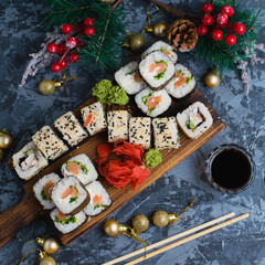 Large set of sushi on a wooden board top view. Rolls with sesame seeds, nori, cheese, red fish, cucumber and eel, ginger, wasabi, soy sauce, bamboo sticks and christmas decor on gray background