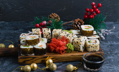 Rolls with sesame seeds, nori, cheese, red fish, cucumber and eel, ginger, wasabi, soy sauce and Christmas decor on a gray background. A large set of sushi on a wooden board.