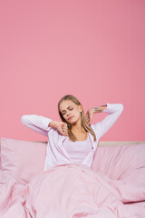 Obraz na płótnie Canvas Young blonde woman in pajamas stretching on bed isolated on pink