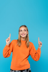 Smiling blonde woman in orange sweatshirt pointing with fingers isolated on blue