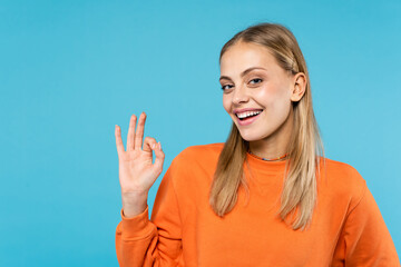 Cheerful woman in orange sweatshirt showing ok gesture isolated on blue