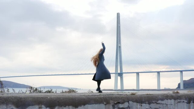 Aerial zooming out view of a woman with long hair dancing in artistic manner. The Russian bridge across Eastern Bosphorus strait and Vladivostok cityscape on the background