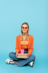Smiling student in sunglasses holding books and paper cup while sitting on blue background