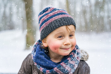 Close up portrait of a little boy with snowflakes in a park in winter. Happy child enjoys the first snow in a forest.