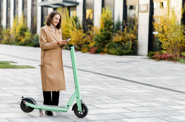 Happy woman in autumn coat using smartphone for renting modern electric scooter with an application, reads qr code with a mobile phone camera. Alternative city transport