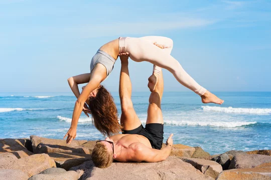 Fit young couple doing acro yoga at spa retreat on sea beach. Active woman  balancing on partner feet, stretching at acroyoga pose. Healthy lifestyle.  People outdoor sport activity on family vacation. Stock Photo