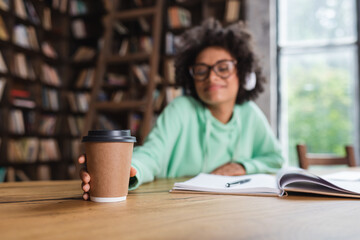 Fototapeta premium blurred african american student in eyeglasses and headphones reaching paper cup