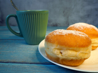 two doughnuts, sprinkled with powdered sugar, on a white plate next to a dark mug of coffee on a blue wooden table. side view