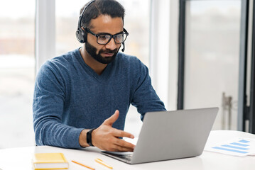 Focused confident male Indian remote consultant wearing headset using laptop computer to virtual...