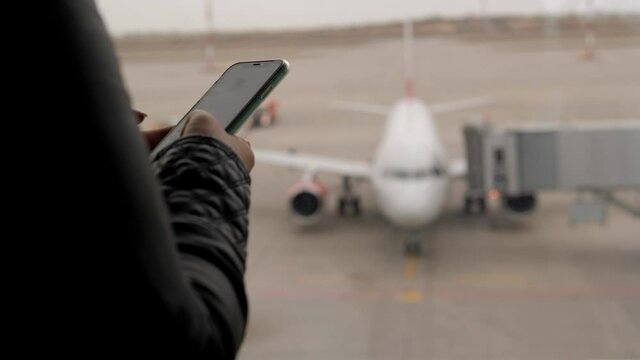 Close-up of a woman holding a phone at the airport near a large window, in the background an airplane sleeve or ladder approaches the plane.