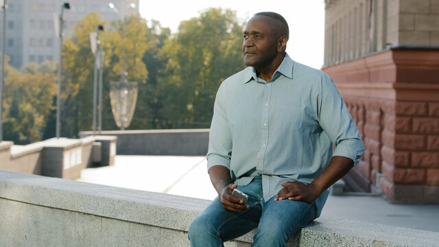 Stressed frustrated elderly African American man of retirement age in casual clothes shirt and jeans sitting outdoor holding phone waiting for client or friend late for meeting, anxiously look aside 